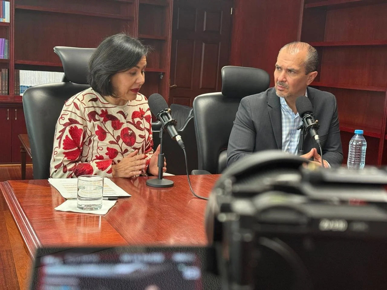 A woman and a man sit at a wooden table with microphones, engaged in a discussion or interview. The woman has black hair, is wearing a cream sweater with red floral embroidery, and the man has light skin, is bald with gray hair on the sides, and is w