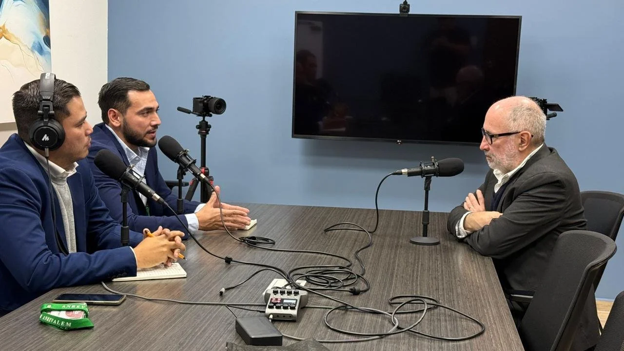 Three men are sitting at a conference table engaged in a discussion, with two microphones set up in front of them and a large screen mounted on the wall behind them.