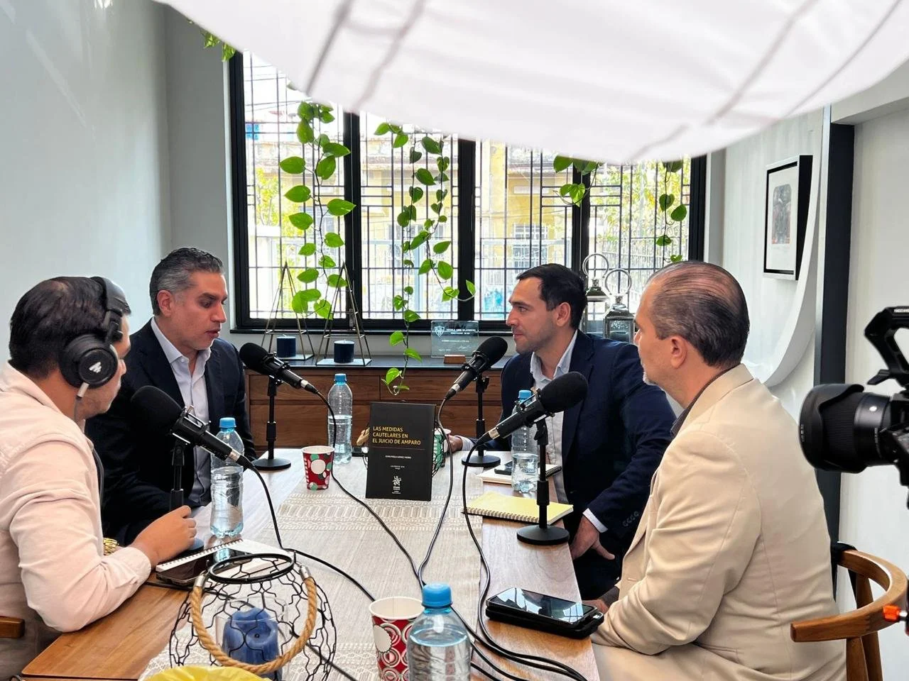 Four men sitting at a table during a podcast recording, with microphones, headphones, water bottles, and a book.