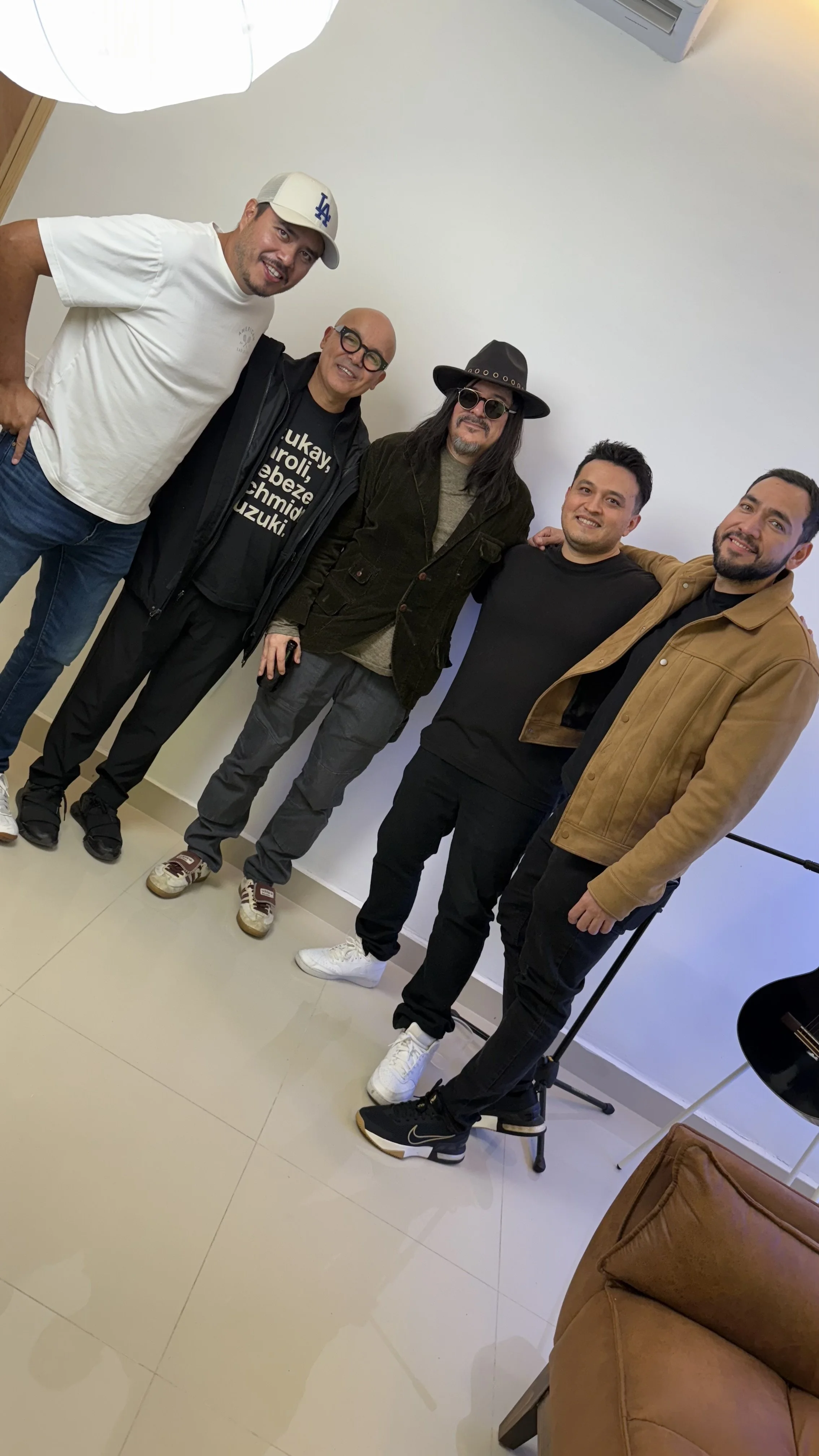 Five men standing together indoors, smiling, with a white wall behind them and a brown couch in the foreground.