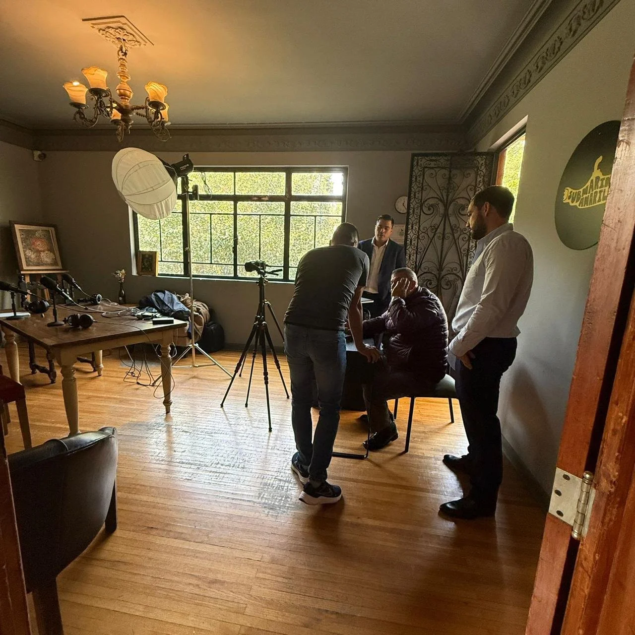 A photo of a small indoor photography or interview setup with four men, some standing and one sitting, in a room with wooden floors, a window, and studio equipment including a large light, umbrellas, and cameras.