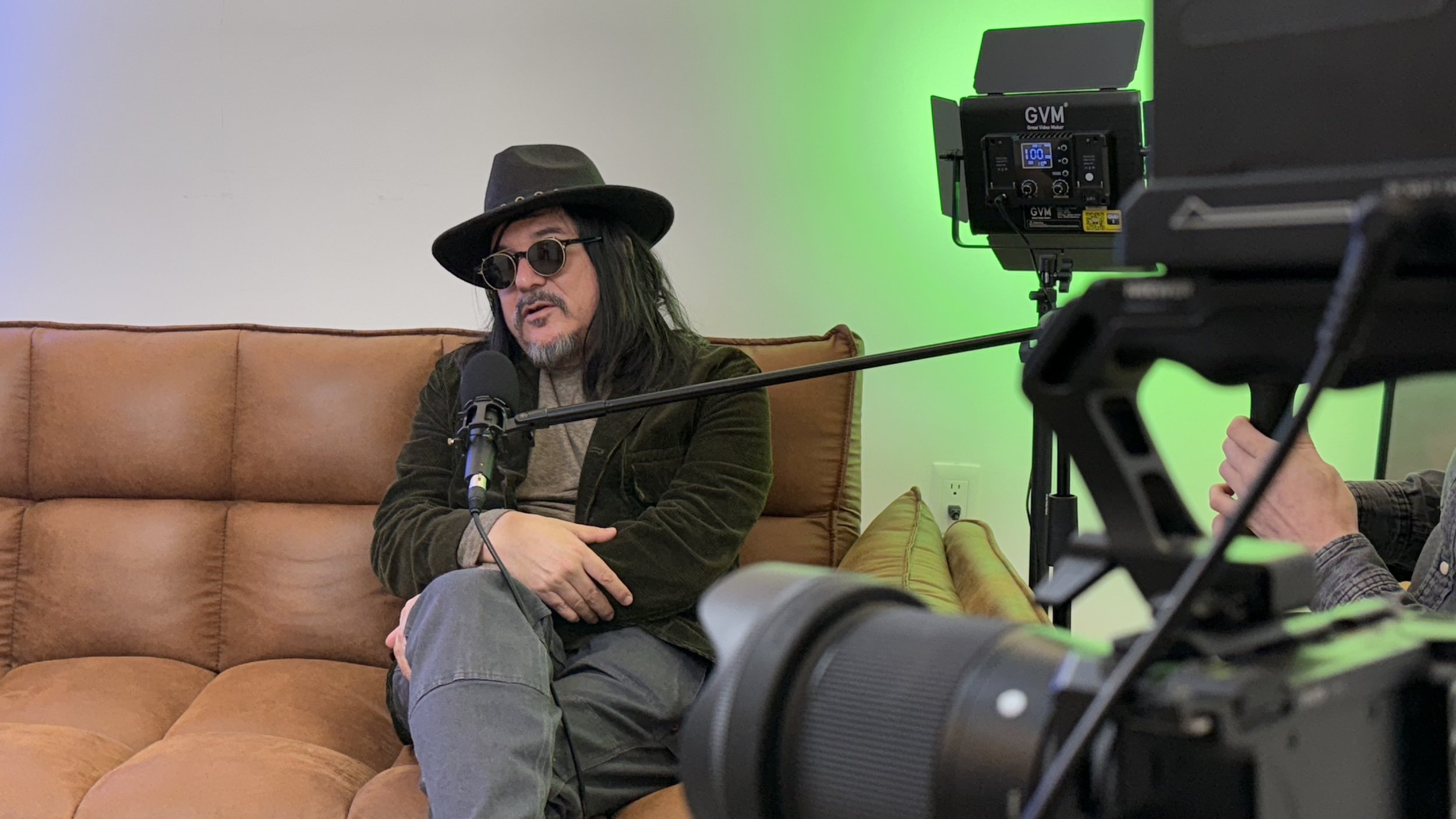 Man wearing sunglasses and a hat sitting on a leather sofa, being filmed with a professional camera and microphone during an interview or recording session, with green lighting in the background.
