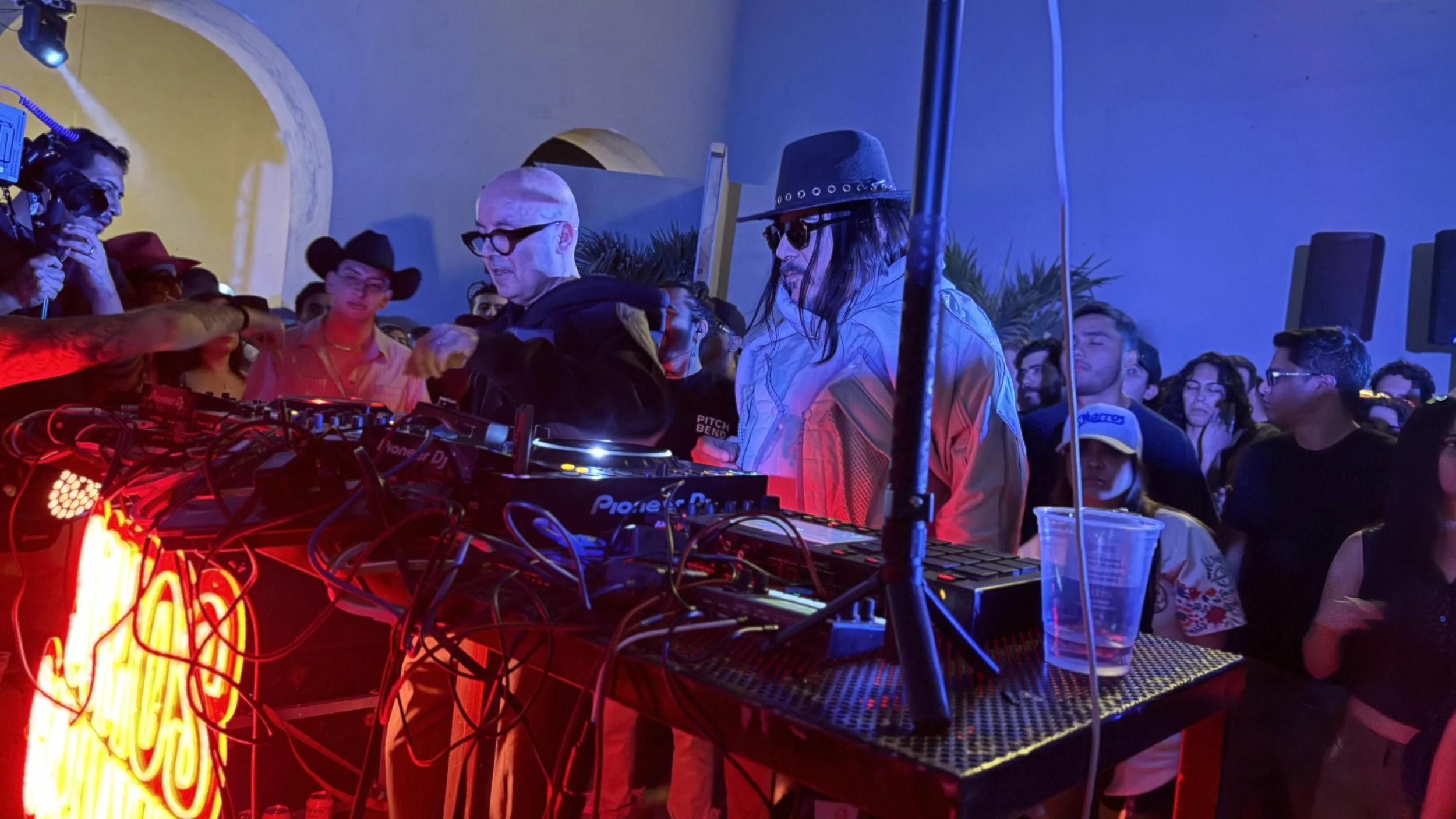 A DJ with glasses and a hoodie playing music at a club with a crowd dancing and socializing, neon lights, and a speaker setup.