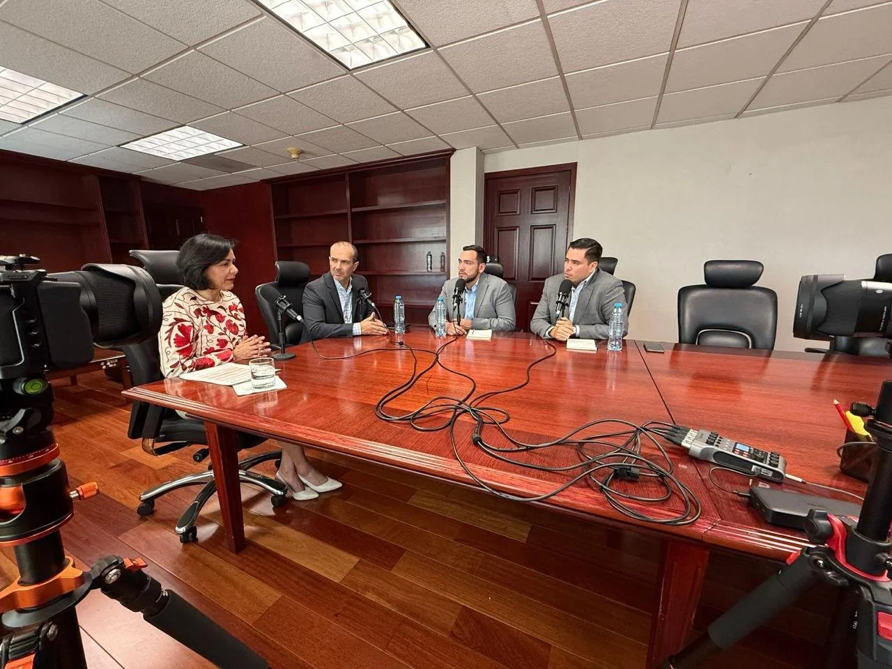 Four people sitting at a large wooden conference table, each with a microphone, participating in a recorded discussion or interview in a professional conference room.