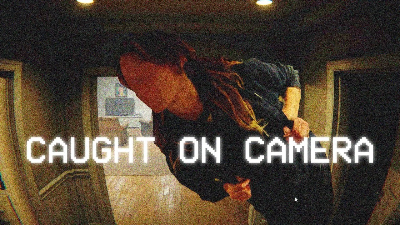 Person with dreadlocks in dark clothing looking down at the camera inside a home.