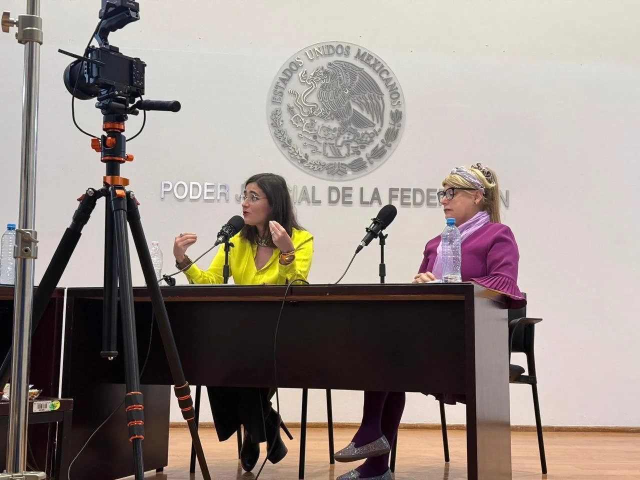 Two women sit at a table in front of a backdrop with the Mexican government emblem and text, speaking into microphones during a press conference or official event.
