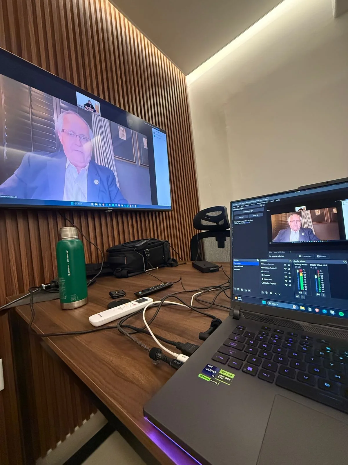 A virtual meeting setup with a large TV screen and a laptop showing a man speaking. The desk has various electronic devices, wires, a water bottle, and a backpack.