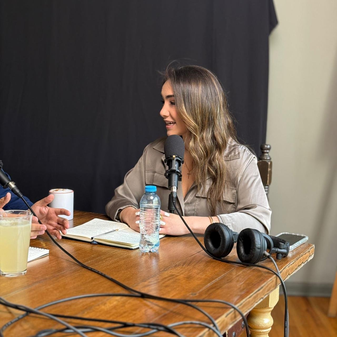 Woman with long wavy brown hair sitting at a wooden table in front of a podcast microphone, engaging in conversation during a podcast recording.