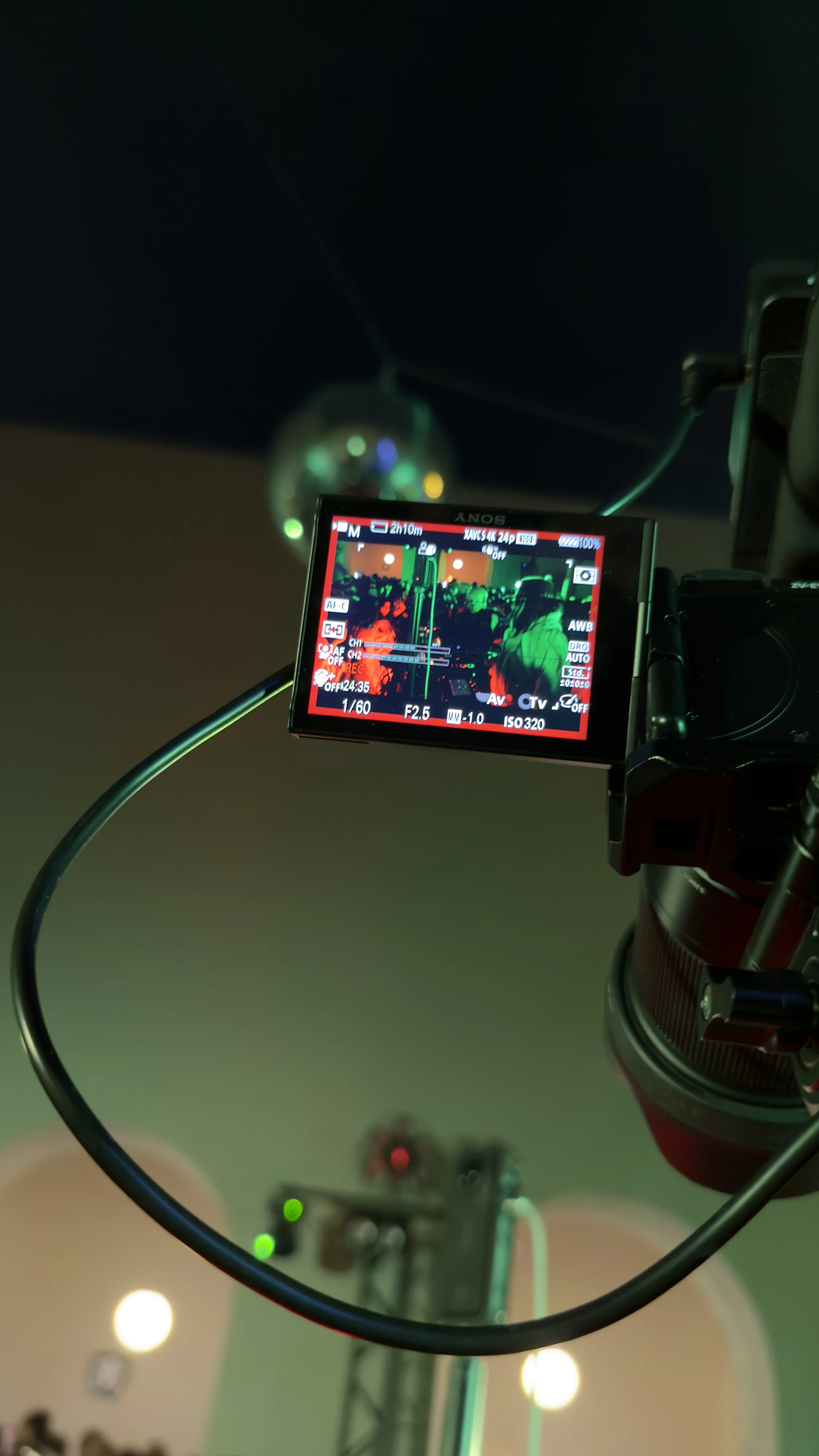 Camera monitor showing a shot of a dance floor with people dancing under colorful lighting, mounted on a tripod with a blurred disco ball in the background.