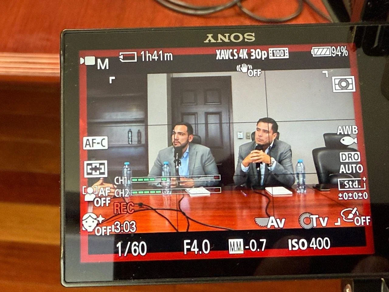 Camera screen displaying two men seated at a table with water bottles during a meeting or presentation.
