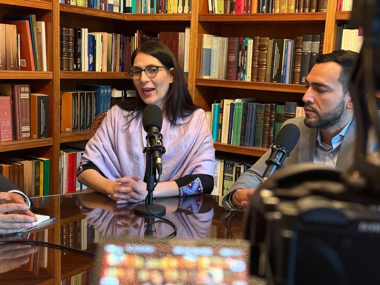 Two people, a woman wearing glasses and a man, sitting at a table with microphones in front of them, in a room with bookshelves filled with books.