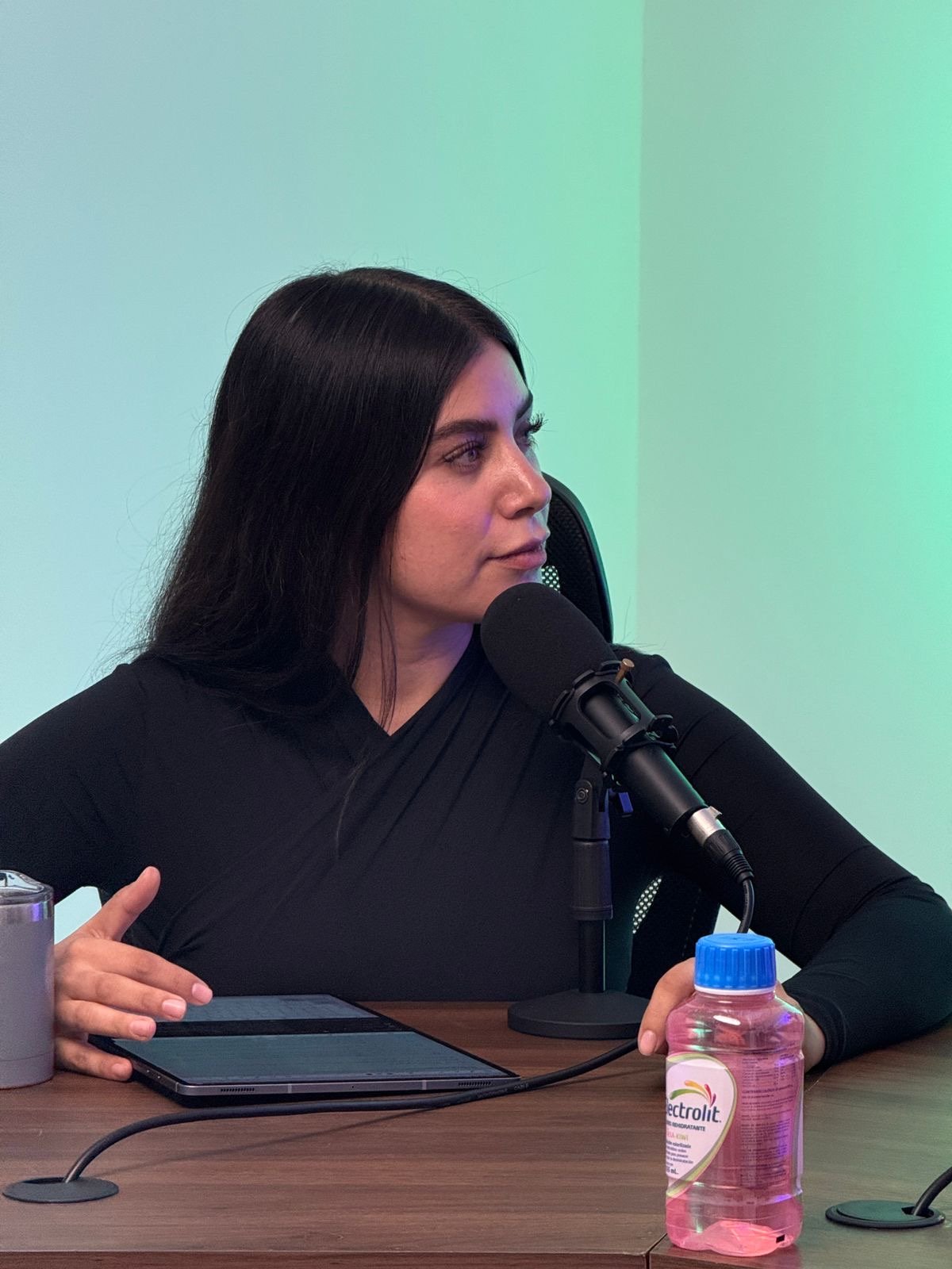A woman with long dark hair and a black top speaking into a microphone during a recording or interview. She is sitting at a table with a tablet, a small bottle of pink hand sanitizer, and a metal tumbler.