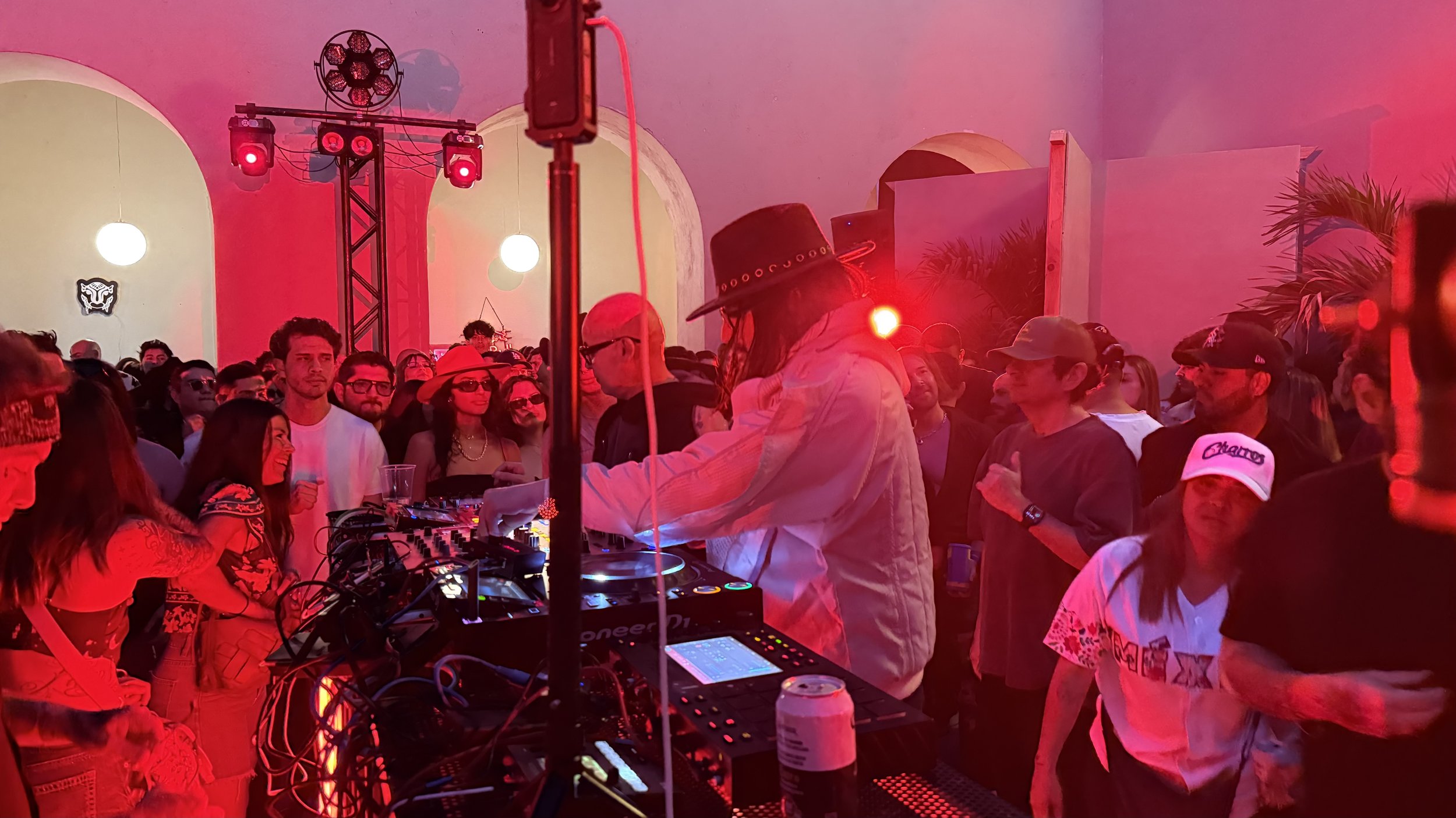 A DJ in a white jacket and a black hat with decorative elements is playing music at a crowded indoor party, with red lighting and various partygoers dancing and socializing.