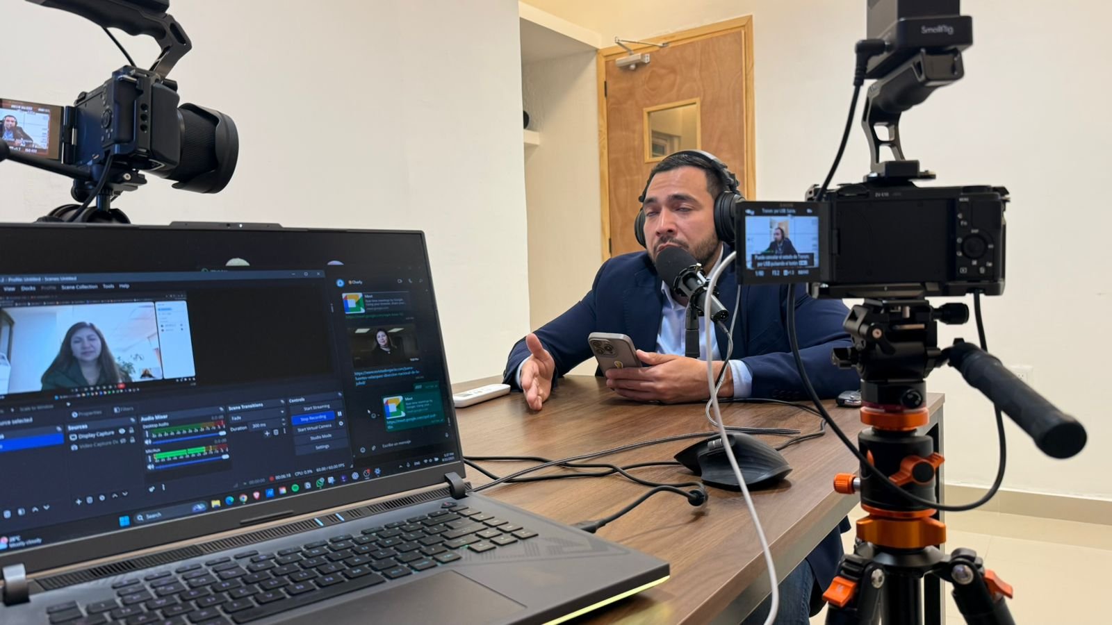 Man in a suit participating in a video interview or recording, seated at a table with a microphone, surrounded by a laptop displaying his video call, a professional camera on a tripod filming him, and another smaller camera on a tripod.