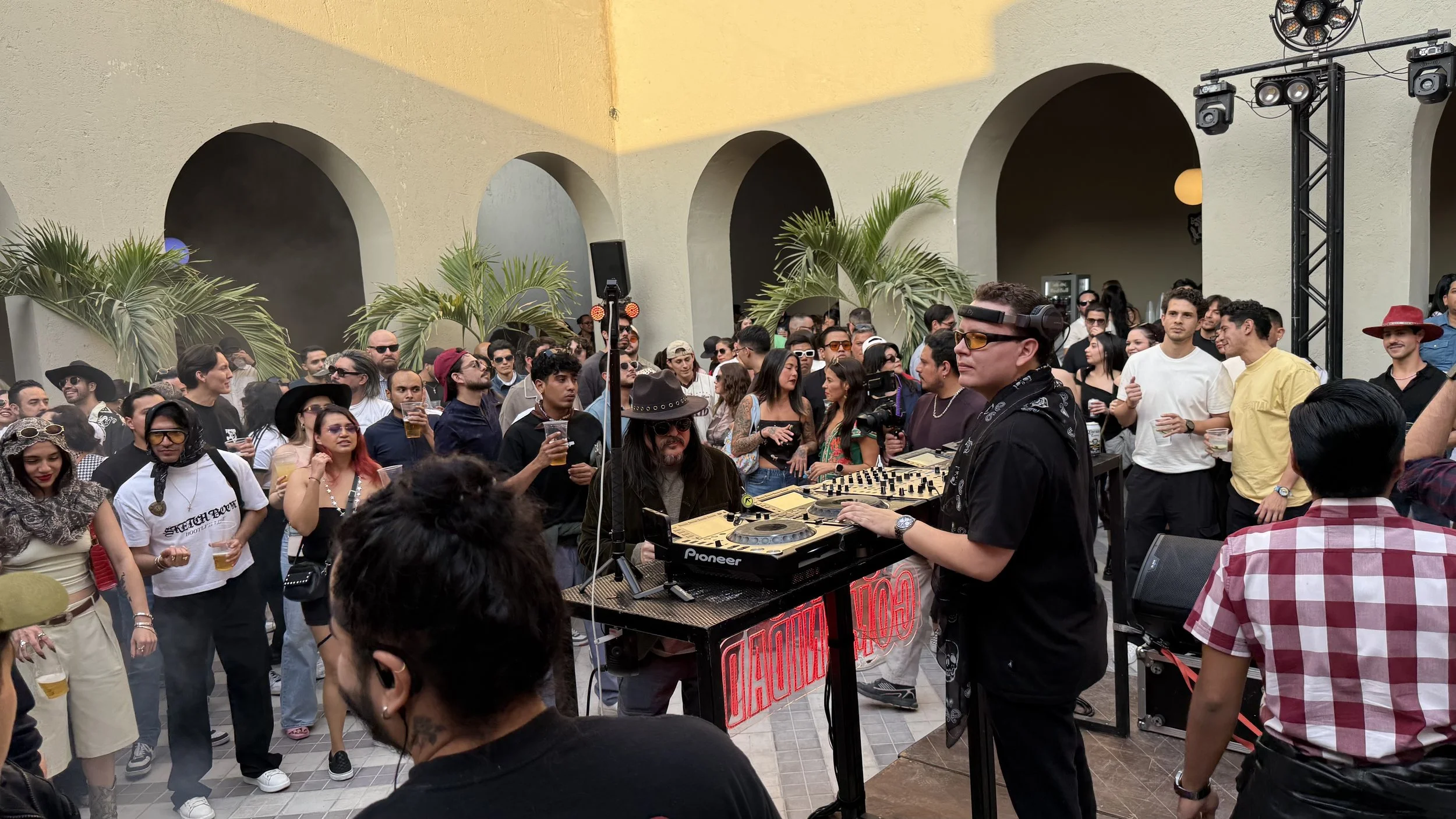Crowd of people dancing and socializing at an outdoor party with a DJ playing music, palm trees, and decorative arches in the background.
