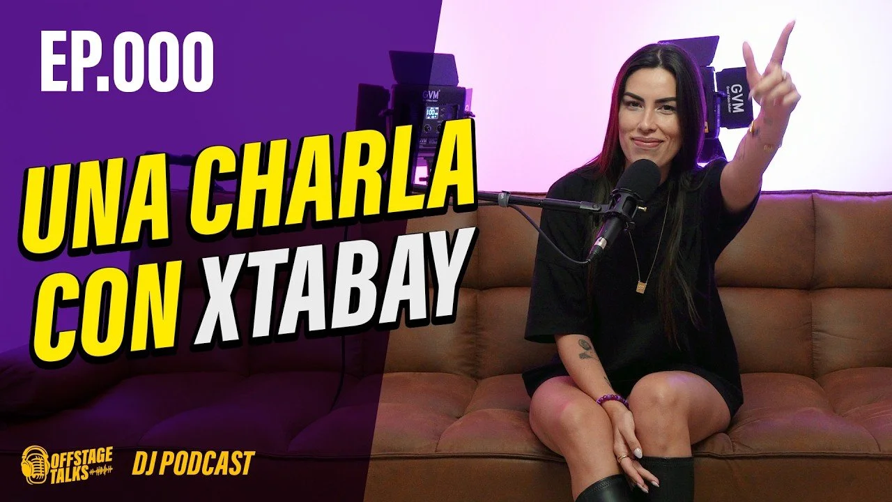 A woman with long dark hair sitting on a beige couch, pointing towards the camera with a smile, in front of a microphone, with studio equipment in the background. The left side has purple and pink lighting and text that reads 'EP. 000 UNA CHARLA CON 