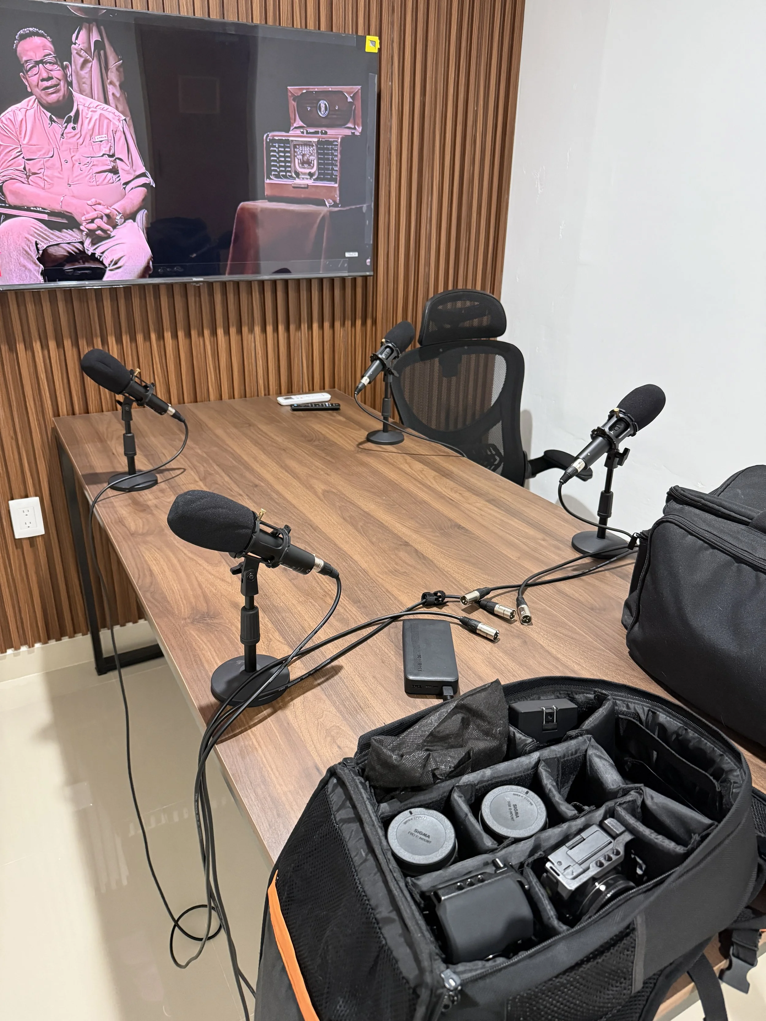 A conference room with a wooden table, four microphones connected to a portable audio recorder, a black ergonomic office chair, and a large screen on a wooden paneled wall displaying an image of a man and vintage radio equipment. Photography equipmen