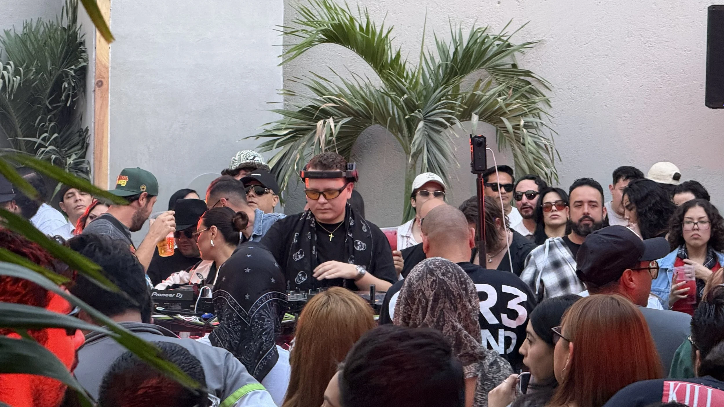 Crowd of people at an outdoor event with a DJ in the center, wearing sunglasses, headphones, and a cross necklace, surrounded by plants and a gray wall.