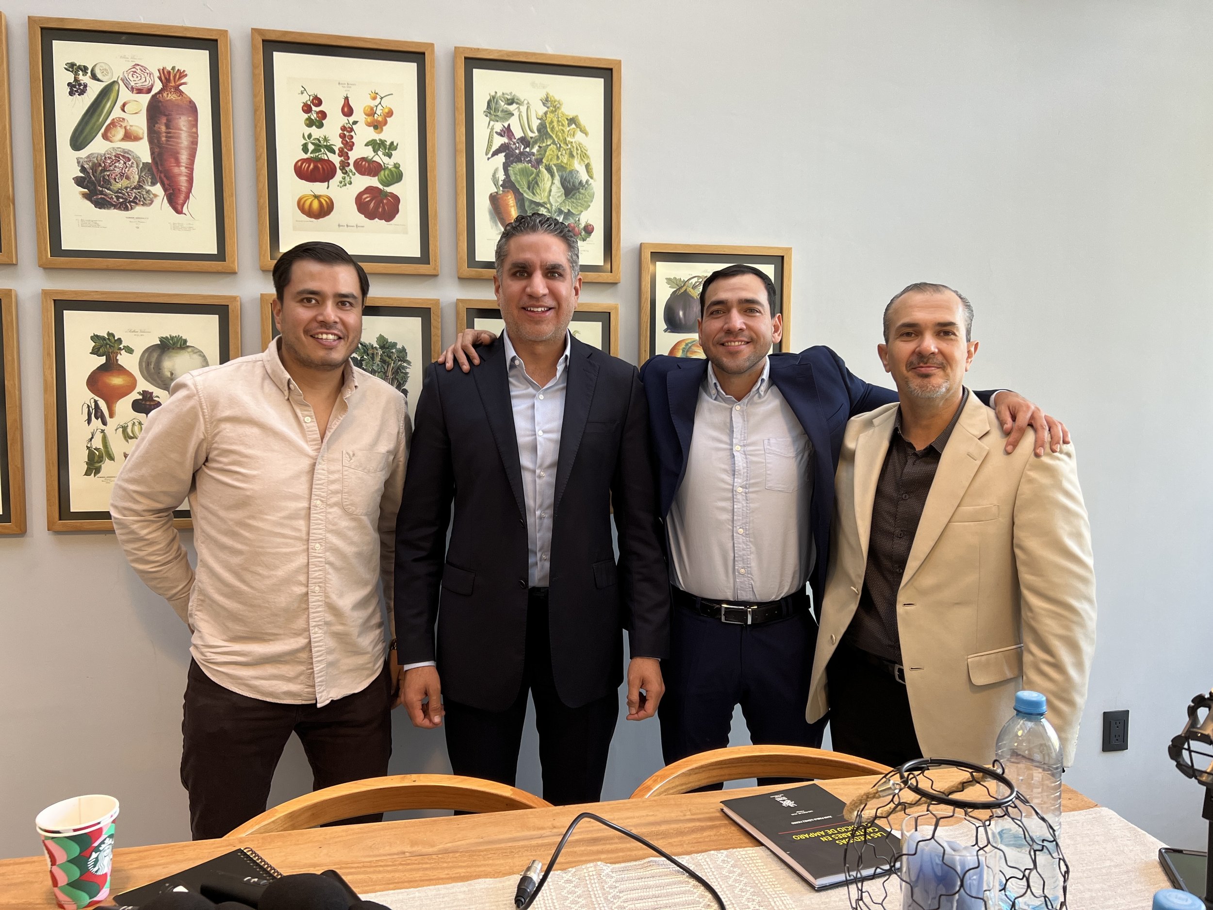 Four men standing side by side with arms around each other indoors, smiling. Behind them is a wall with framed botanical prints of vegetables, tomatoes, and other plants. In front of them is a wooden table with a notepad, a black object, a water bott