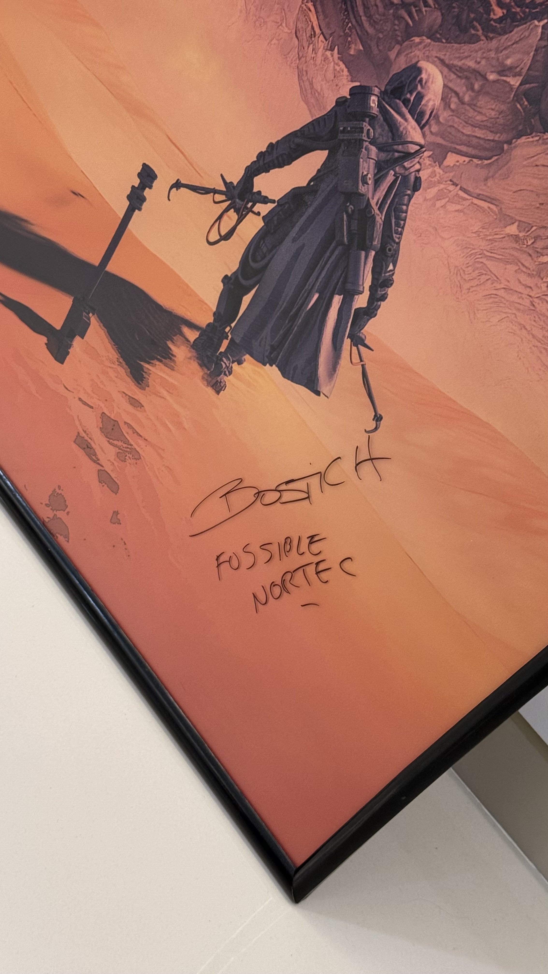 A framed print of a futuristic soldier in a desert landscape with mountain in the background and the words 'BOSTICK' and 'FOSSIBLE NORTEC' handwritten on the glass.