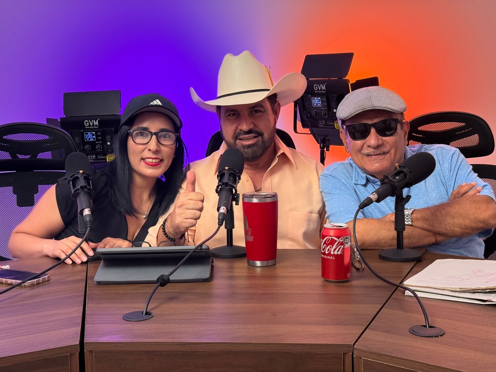 Three people sitting at a table with microphones, recording equipment, and drinks, against a colorful gradient background.