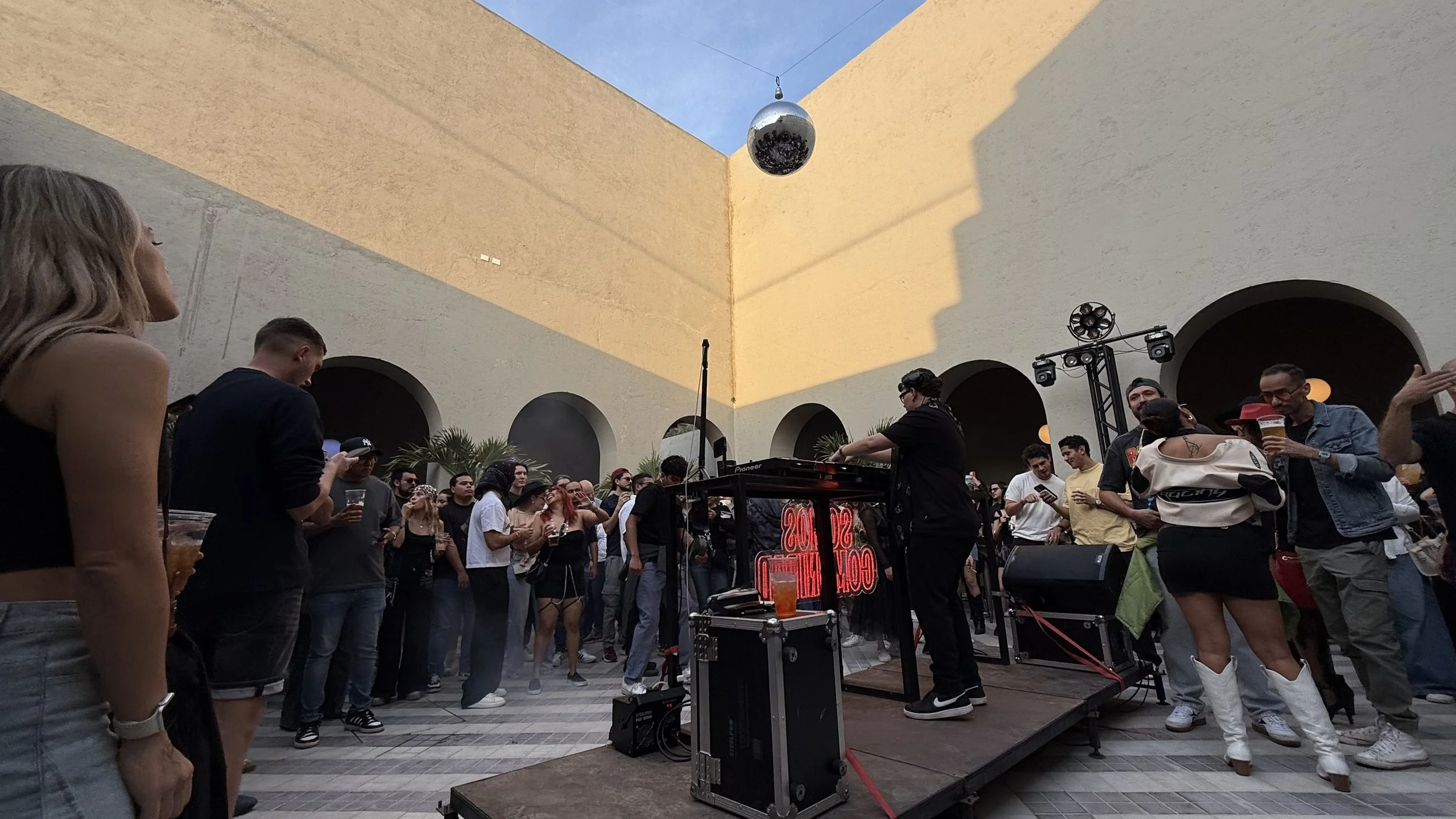 People gathered at an outdoor music event with a DJ performing on a small stage. A disco ball hangs overhead, and the audience is watching and dancing.
