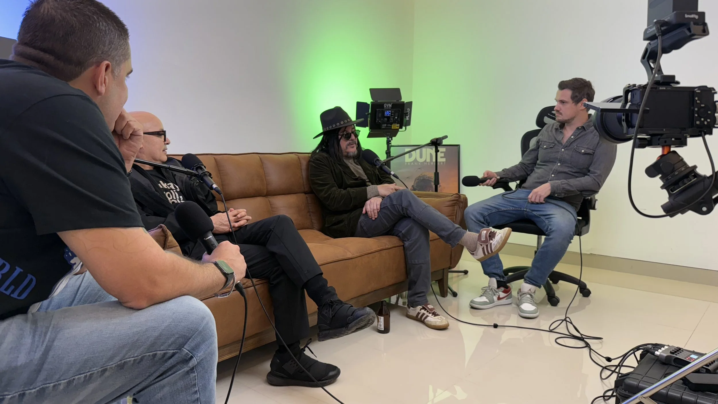 Four people sitting on a brown leather couch and a chair in a recording studio, engaged in an interview. One person is wearing a cowboy hat and sunglasses, speaking into a microphone, while another is holding a microphone and sitting in a chair. Two 