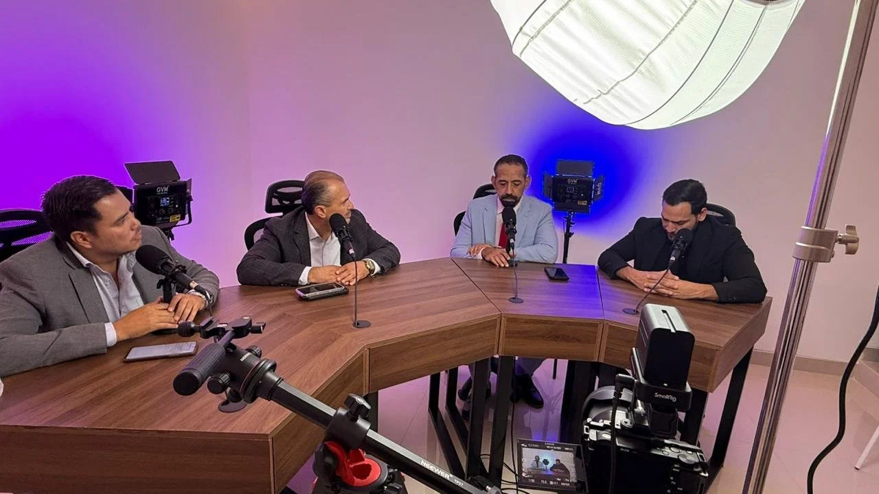 Four men sitting at a curved wooden table with microphones during a podcast or panel discussion, with studio lighting and equipment visible.