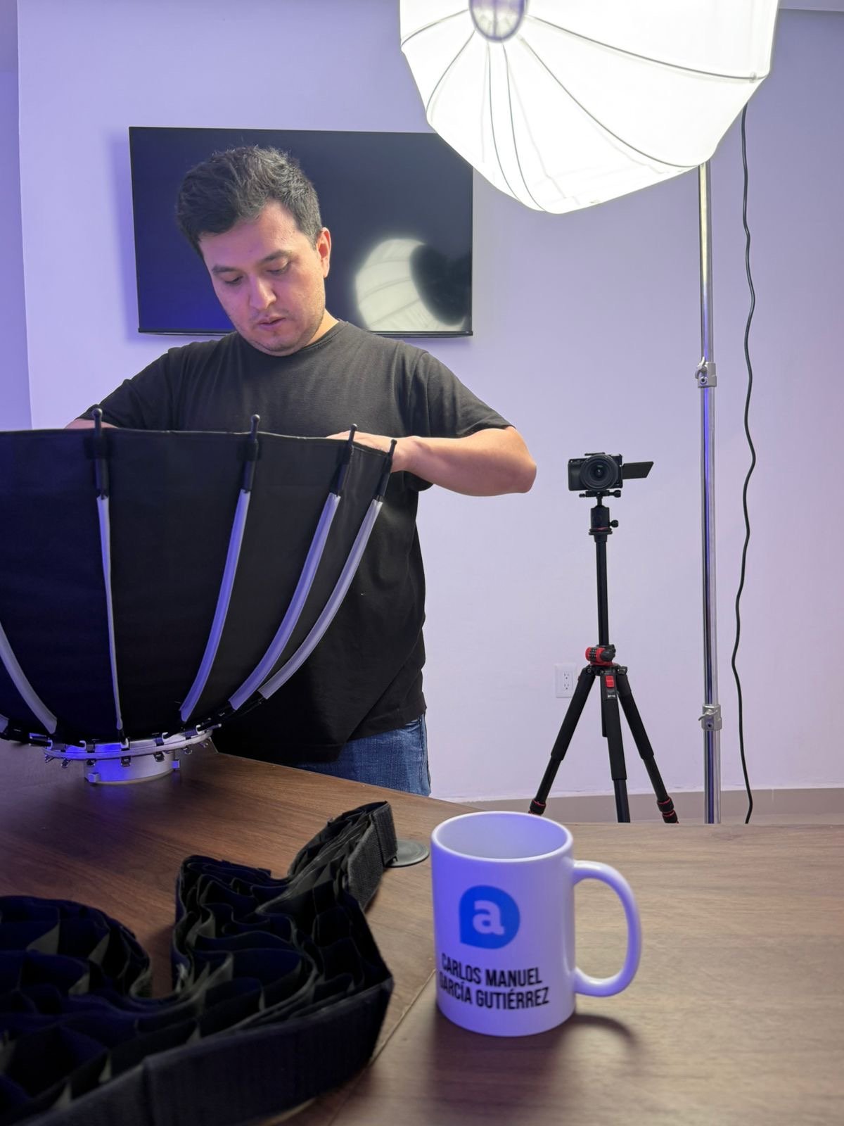 Man setting up photography or video equipment in a studio with a large softbox light overhead, a camera on tripod, a TV on the wall, and objects including a white mug with text on the table.