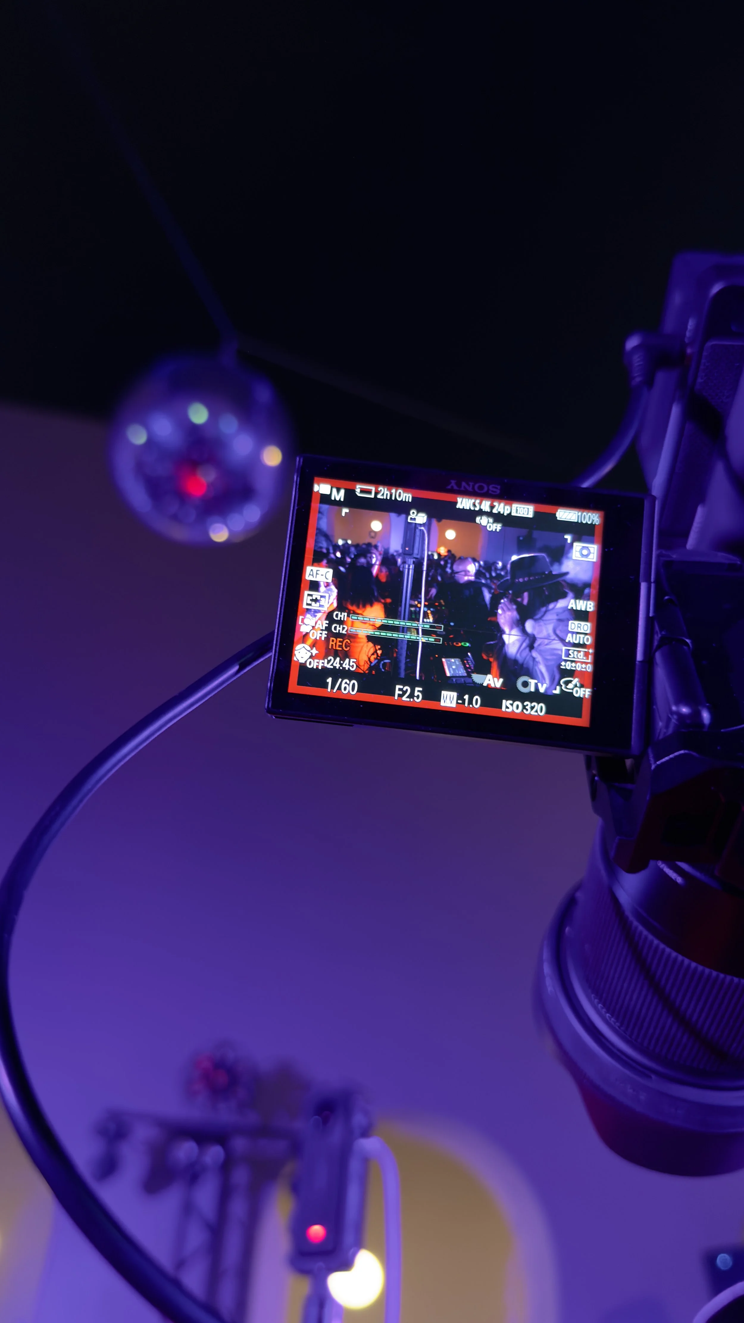 A professional video camera on a tripod shoots at an event with purple and orange lighting, capturing a crowd of people.