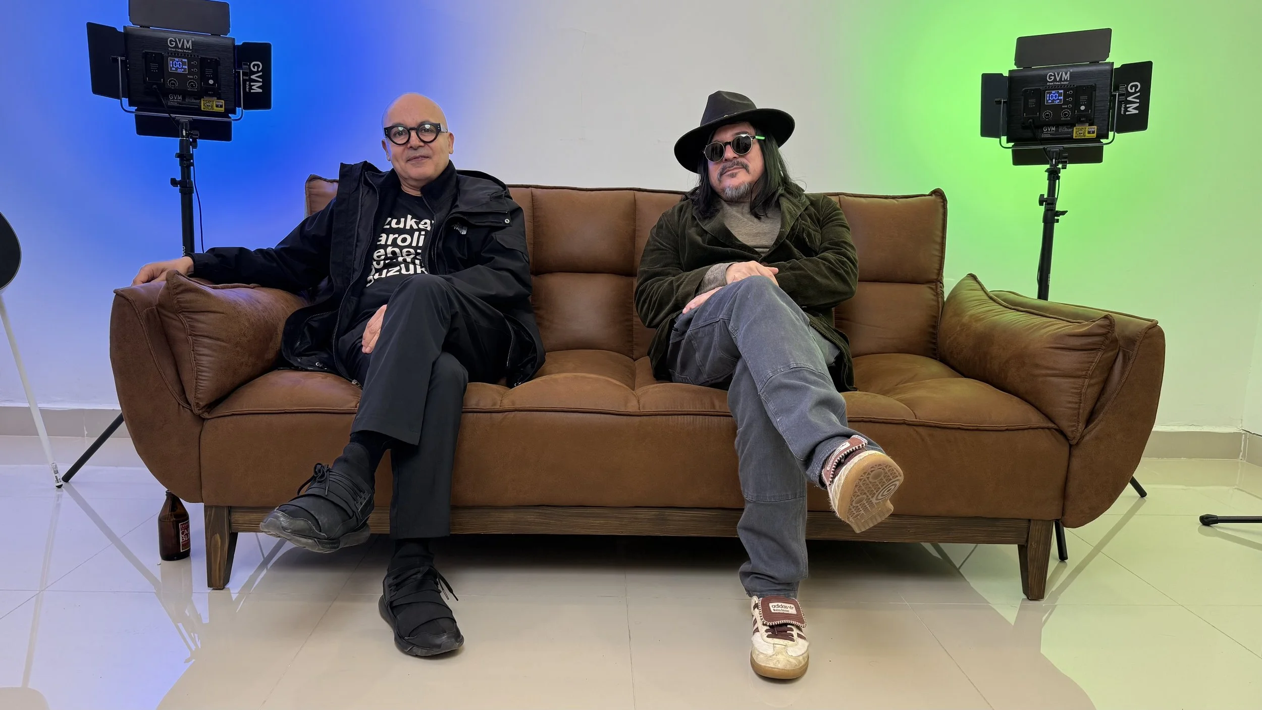 Two men sitting on a brown leather sofa in front of a white wall with green and blue lighting. They are wearing casual and artistic clothing, with one wearing glasses and a hat and the other younger person with sunglasses.