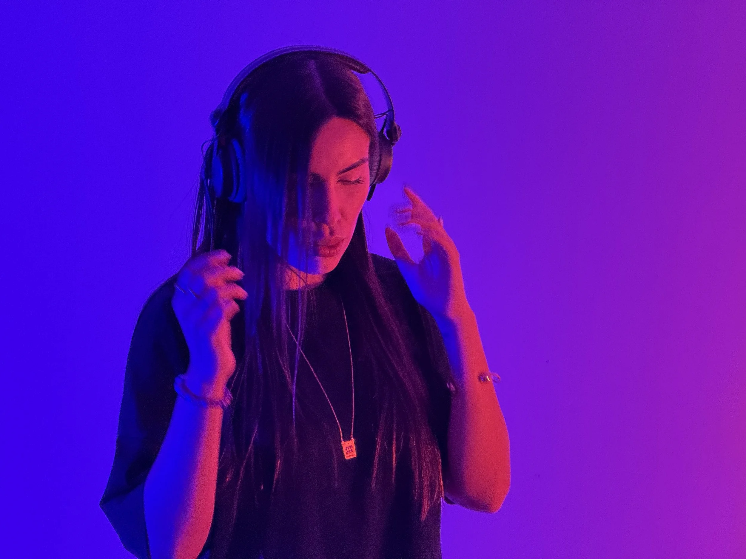 A young woman with long hair listening to music through large headphones, eyes closed, reacting to the sound, illuminated by vibrant purple and pink lighting.