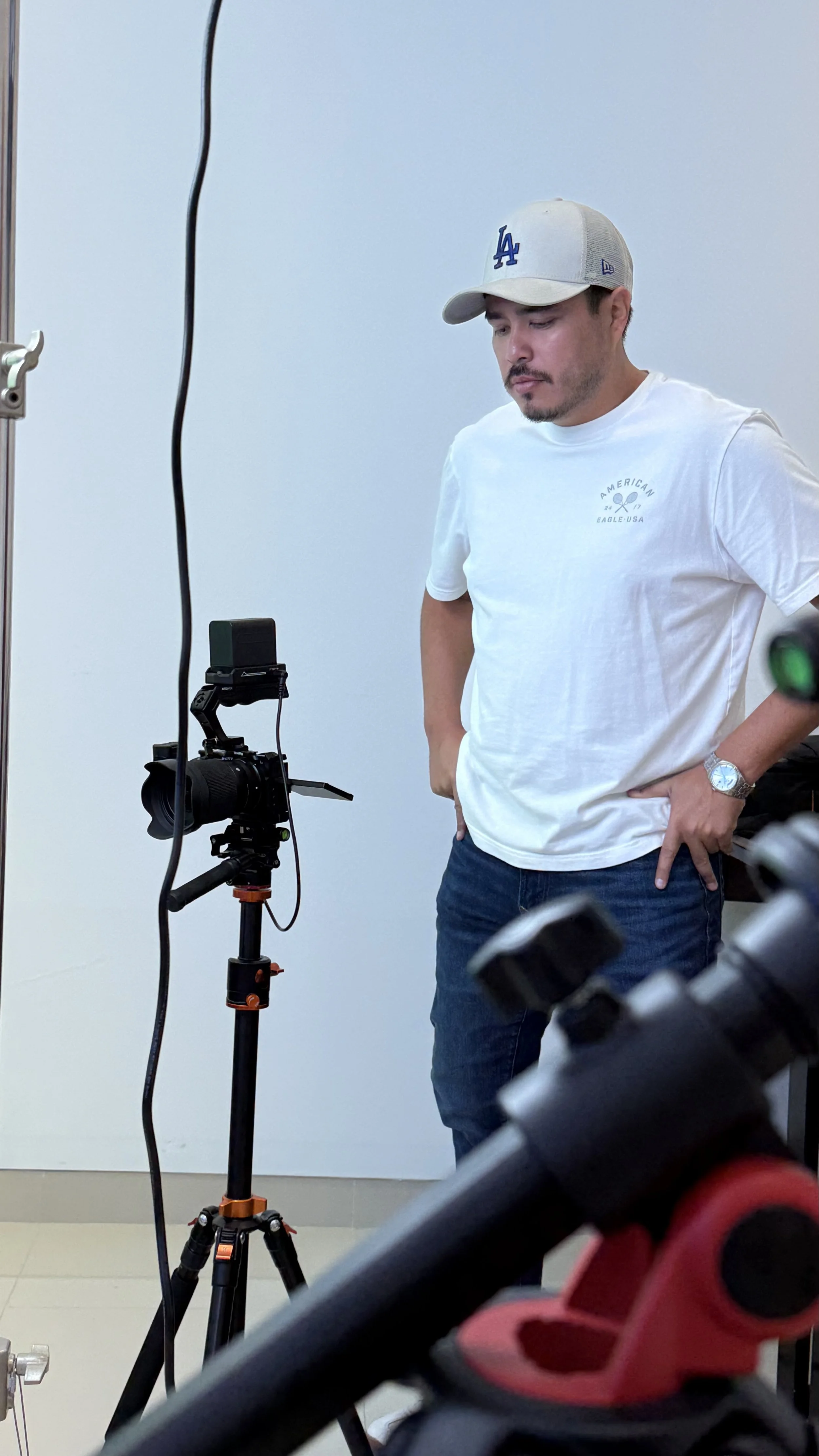 Man in a white t-shirt and gray Los Angeles baseball cap standing in front of a camera on a tripod in an indoor setting.