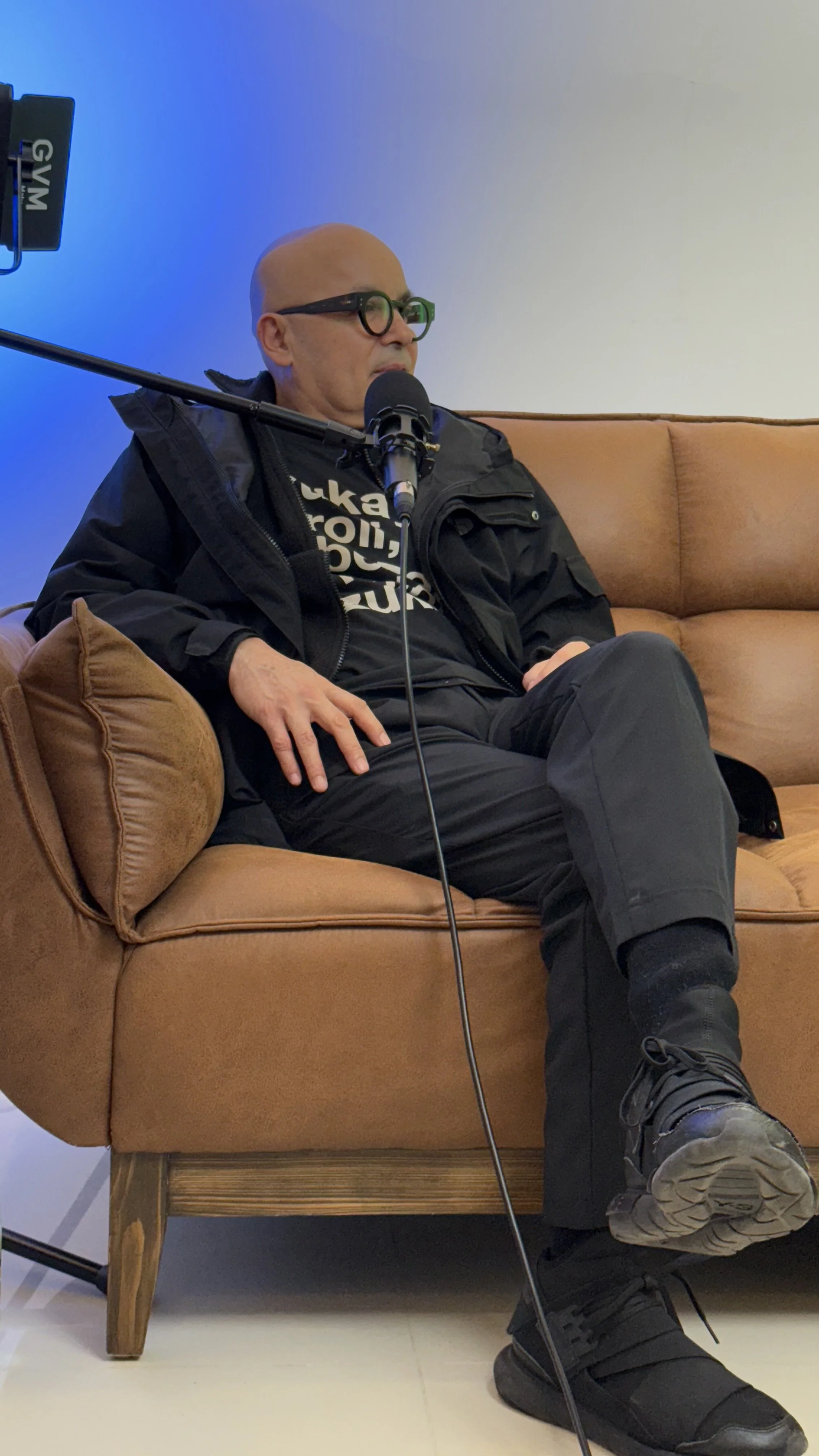 A man sitting on a tan couch speaking into a microphone during a podcast or interview, with a blue gradient background and professional lighting.