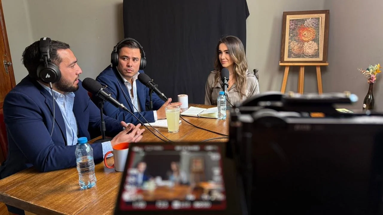 Three people participating in a podcast recording, sitting at a wooden table with microphones, headphones, beverages, and notes, in a room with framed artwork and a flower vase in the background.