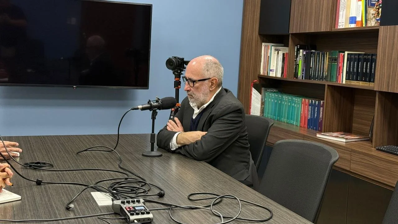 A man with glasses and a beard and bald head, wearing a gray suit and white shirt, sits at a conference table with arms crossed, preparing to speak into a microphone during an interview or recording session in a room with a large television screen an
