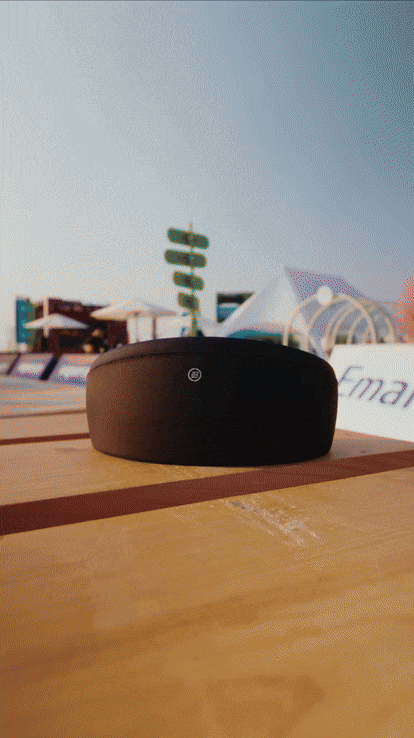 A black, oval-shaped electronic device with a small circular button on top, placed on a wooden surface outdoors with a cloudy sky, a green sign, and tents or booths in the background.