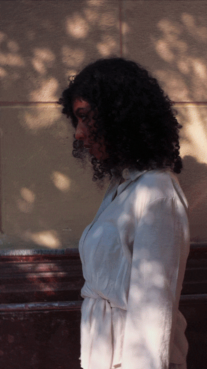 Side view of a woman with curly hair wearing a white coat, standing outdoors with sunlight and shadow patterns on a beige wall behind her.
