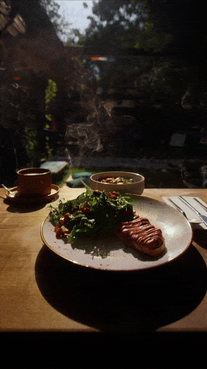 A plate with grilled steak and salad, a cup of coffee, and a bowl of soup on a wooden table near a window with a view of trees and a waterfall.