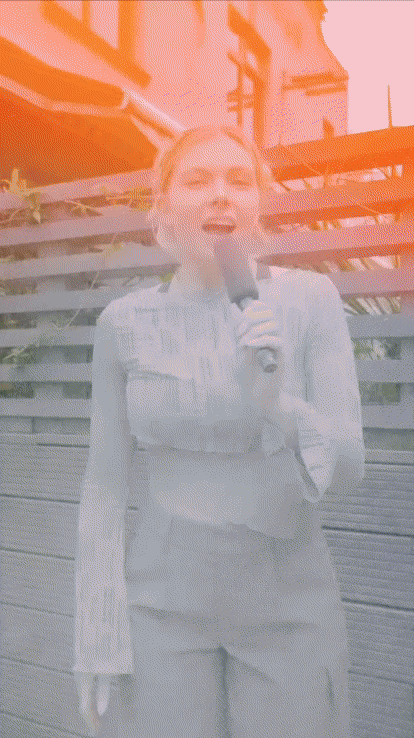 A woman with light-colored hair holding a microphone and speaking outdoors in front of a wooden fence with tree branches and a building in the background.
