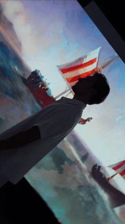 A person standing near a body of water with boats, one of which has an American flag. The person looks toward the water during sunset or sunrise.