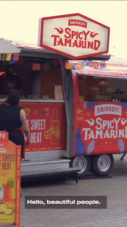 A food stand with a sign that reads 'Smirnoff Spicy Tamarind,' featuring a red and blue color scheme and decorative banners, with a customer standing nearby.