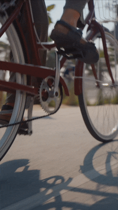 Close-up of a person riding a red bicycle, showing their foot on the pedal and the bicycle's chain and crankset, with a sidewalk and blurred background.