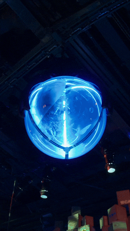 A blue neon light in the shape of a sphere hanging from the ceiling in a dark indoor space.