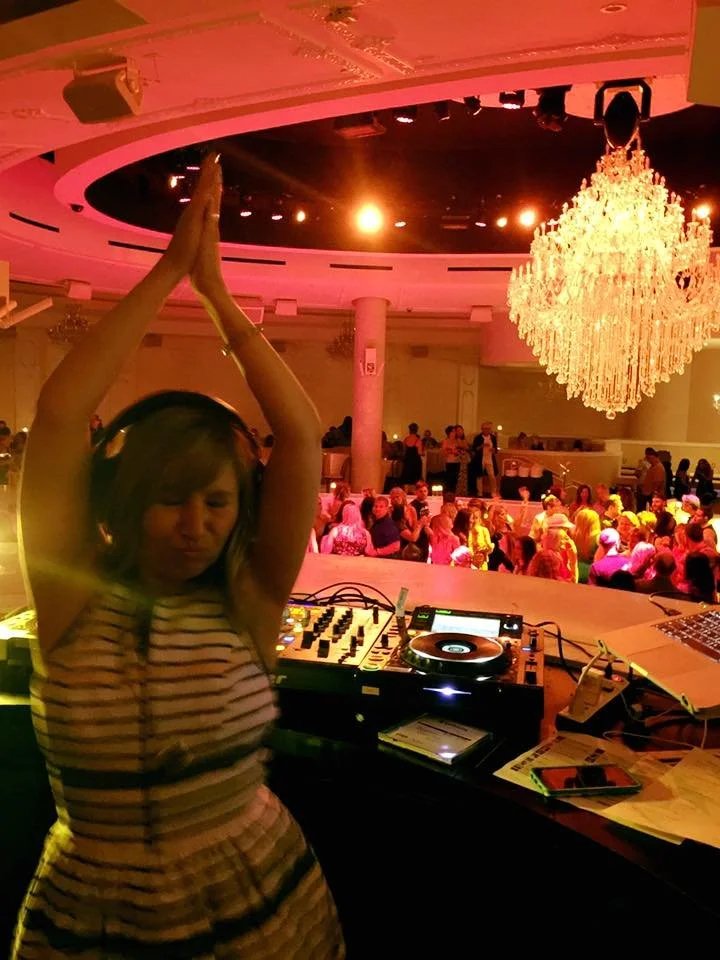 A woman with headphones dancing and raising her arms above her head at a party or event, with a crowded dance floor and a large chandelier in the background.