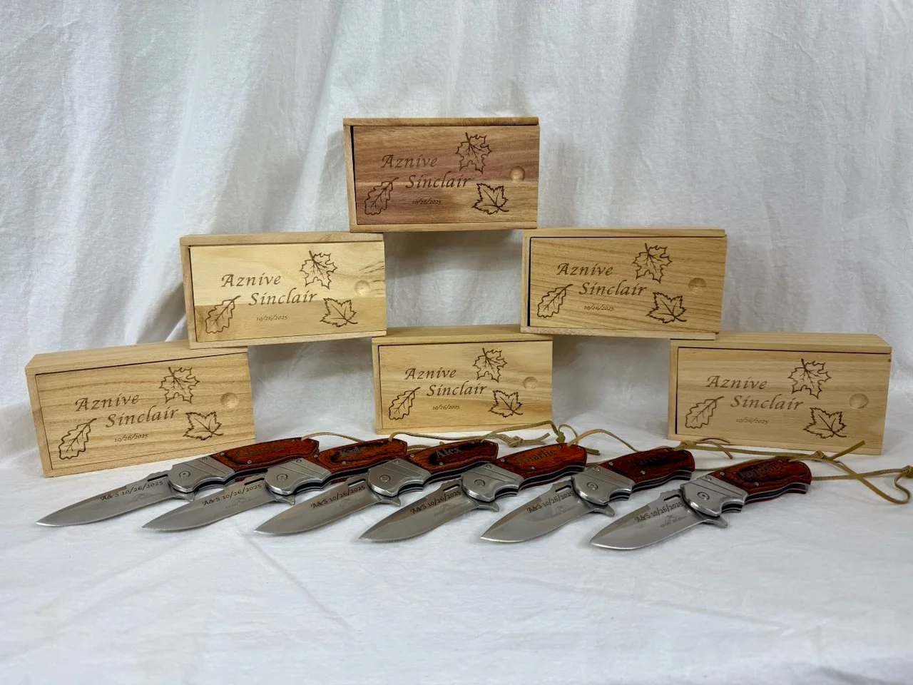 Six pocket knives with wooden handles and metal blades are laid out in front of five wooden boxes with engraved text and leaf designs, set against a white background.