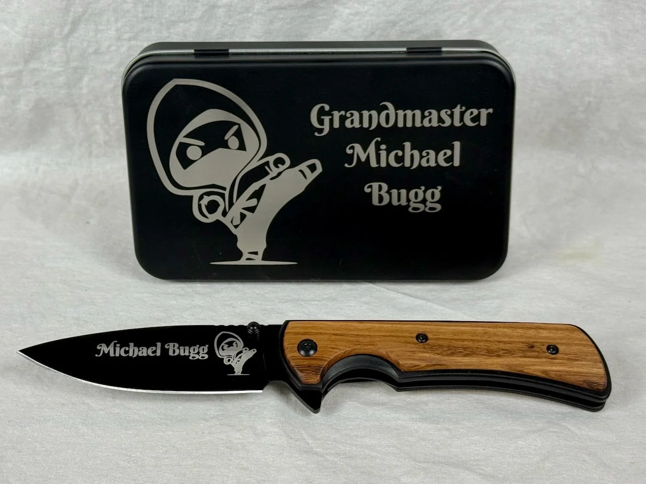 A black folding knife with a wooden handle and a black blade featuring a graphic of a ninja and the text "Michael Bugg." A metal tin box behind the knife labeled "Grandmaster Michael Bugg" with a ninja graphic.