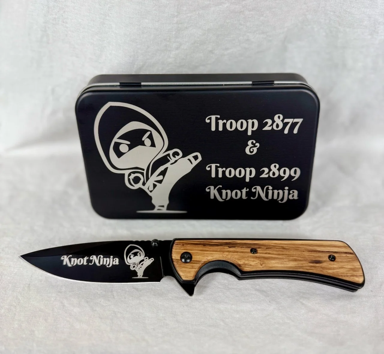 A folding knife with a wooden handle and black blade, engraved with 'Knot Ninja', placed in front of a black metal box that features a ninja illustration and text reading 'Troop 2877 & Troop 2899 Knot Ninja'.