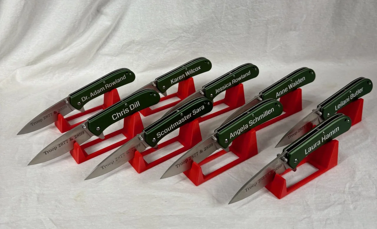 Collection of small knives with green handles and silver blades, each bearing a different name, displayed on red stands.