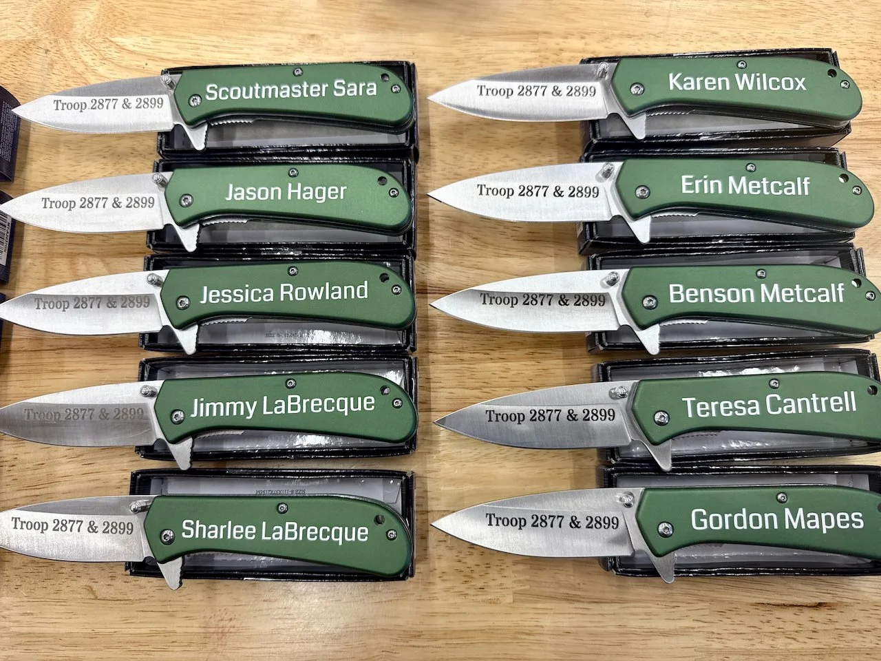 Ten military-style pocket knives with green handles, each inscribed with a name and Troop 2877 & 2899, displayed on a wooden surface. The knives are arranged in two columns, five in each, with the names Jessica Rowland, Jason Hager, Scoutmaster Sara,